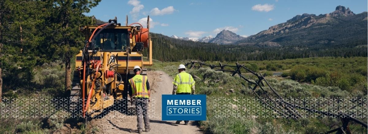 Hero image: two lineworkers walking on trail, back facing, yellow tractor on left side. Mountains in distance 